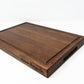 Medium size wood cutting board, made of dark walnut, with a juice groove and side handles.