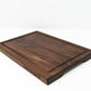 Small wood cutting board, made of dark Walnut, with a juice groove and side handles.